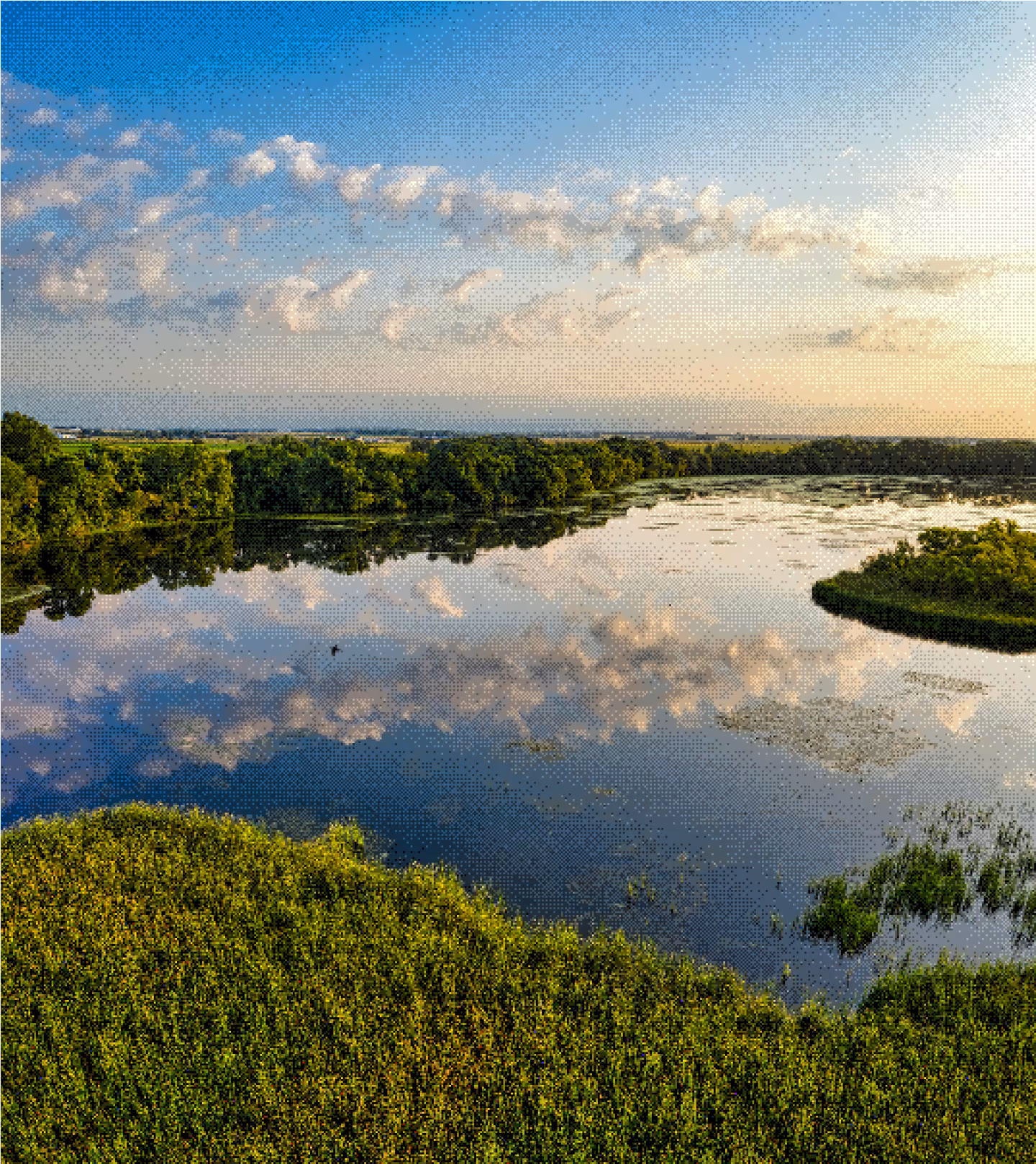 Calm lake reflecting clouds bordered by green trees and vegetation under blue sky at sunset.