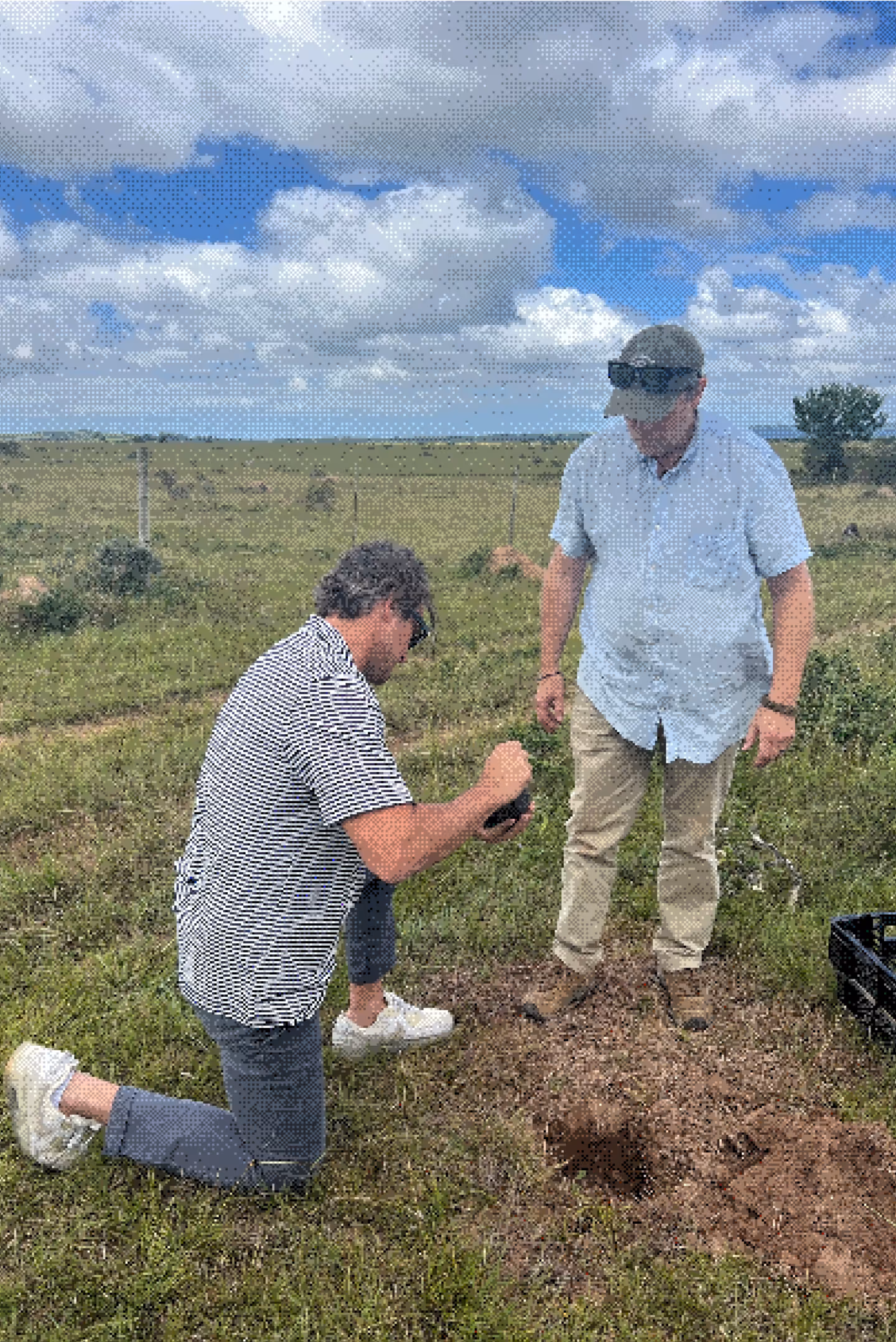 Two men examining soil in a rural field with one kneeling and the other standing nearby.