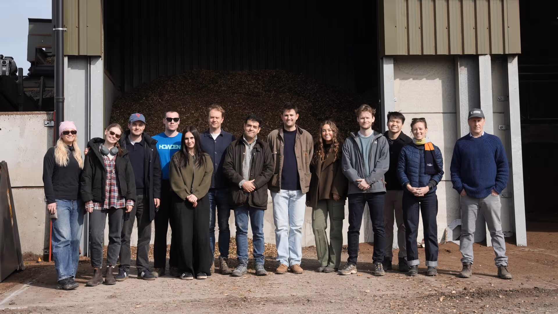 Group of 13 people standing outdoors in front of a large pile of shredded biomass inside a shed.