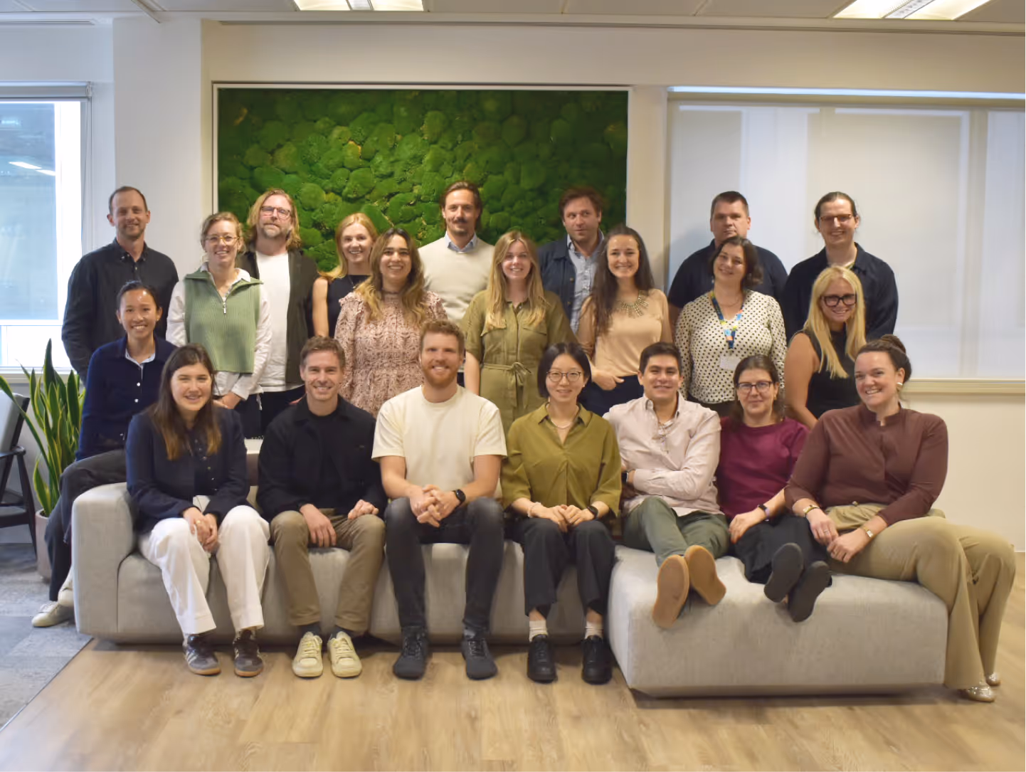 Group of 21 smiling people posing indoors on and behind a light-gray couch with a green wall art behind.