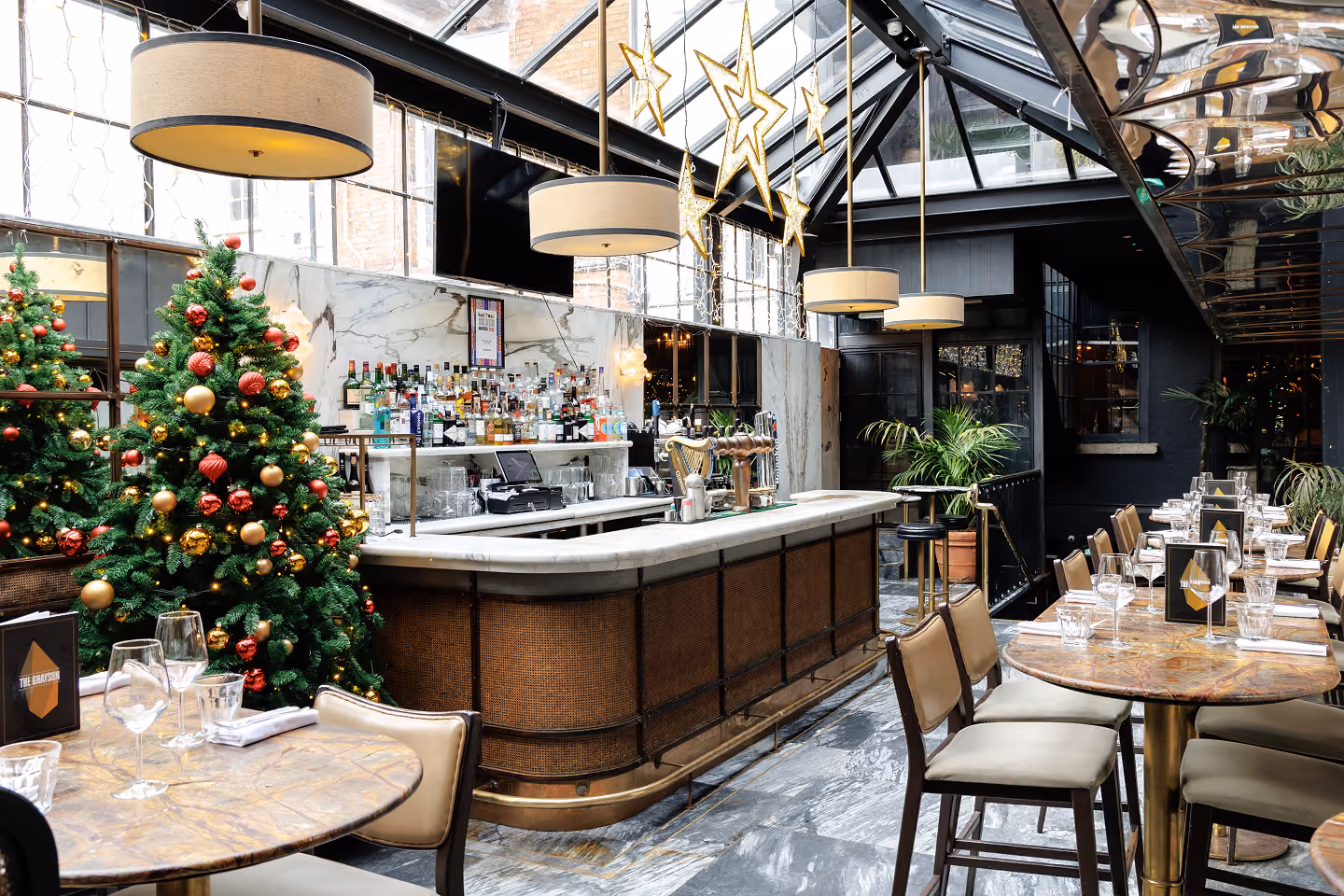 The glass roofed bar at The Grayson at Christmas, with decorated trees and gold stars above set tables