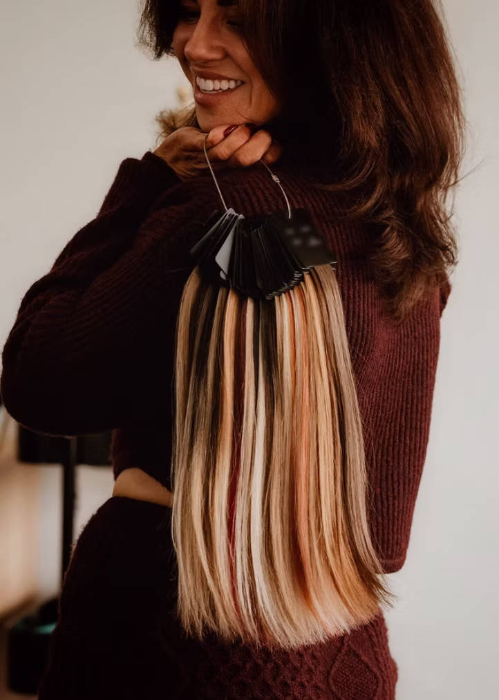 Woman holding a color ring of hair extension samples showing various shades of blonde and brown.
