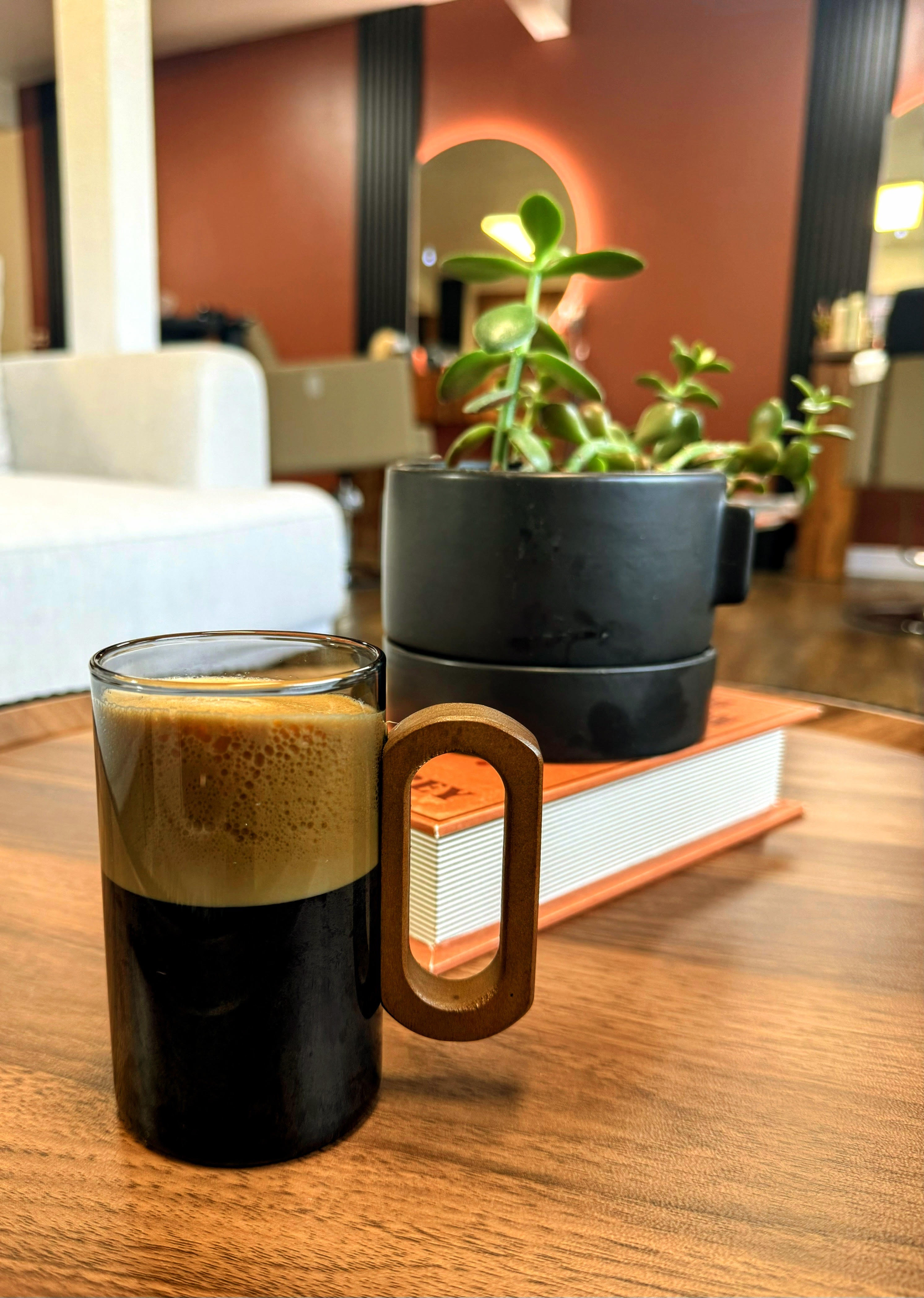 Glass cup of black coffee with foam beside a plant in a black pot on a wooden table.