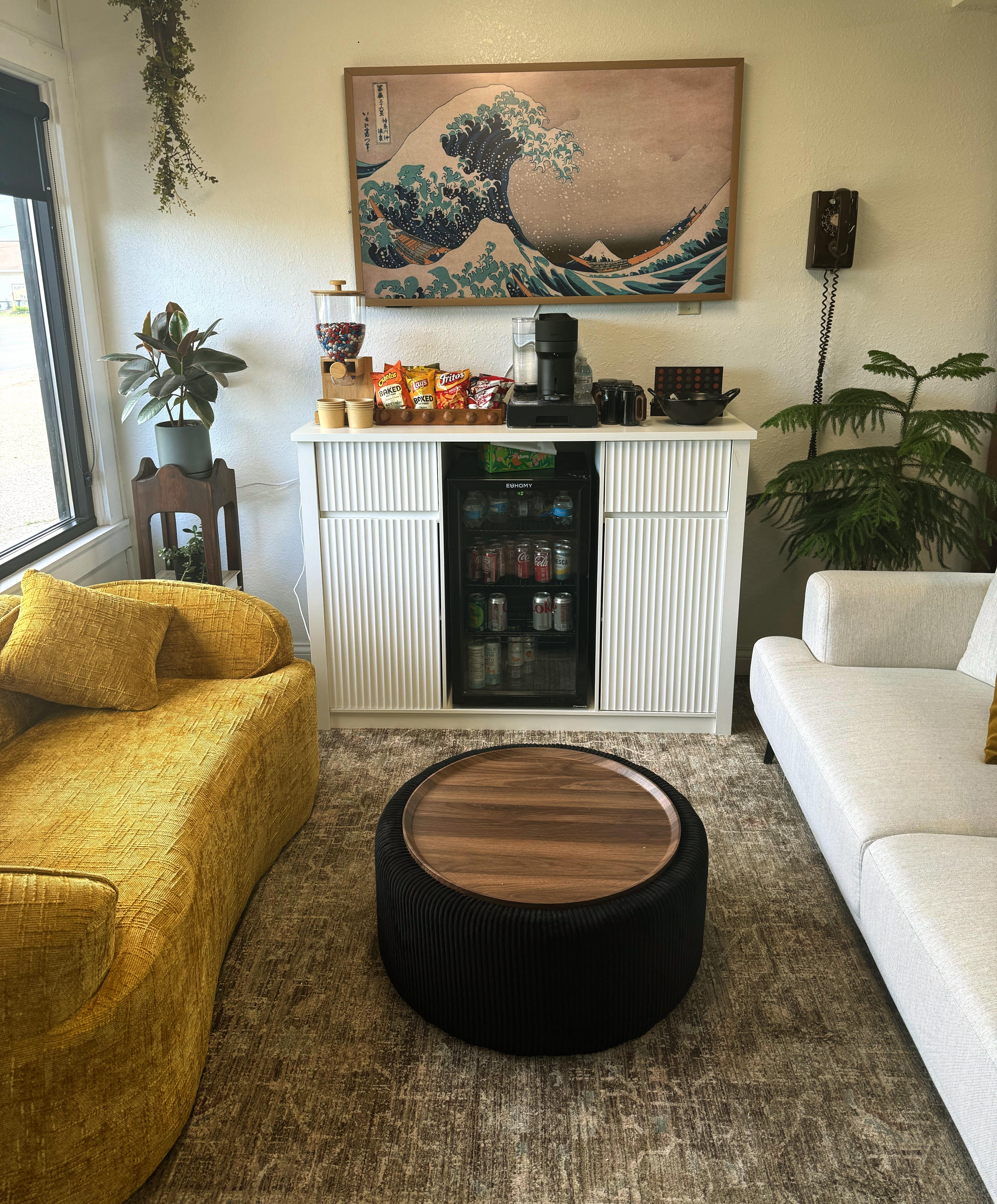 Lounge area with yellow and white sofas, a black round table, a white cabinet with snacks, drinks, and art above.