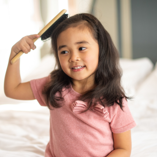 Young girl smiling while brushing her long dark hair with a wooden hairbrush on a bed.