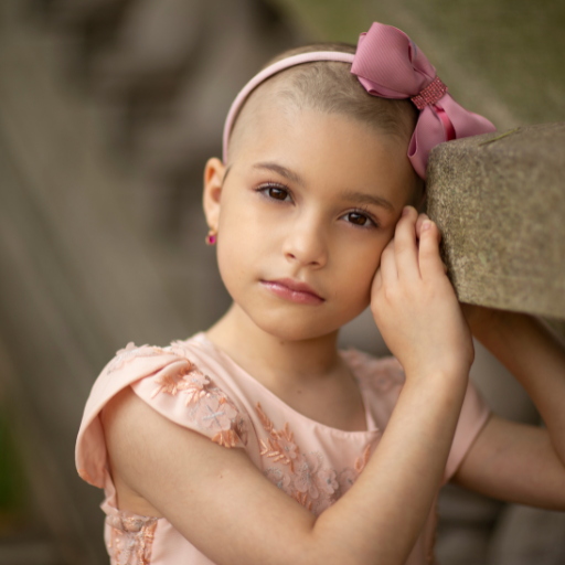 Young girl with short hair and pink bow, wearing a peach dress, resting her head on a stone surface.