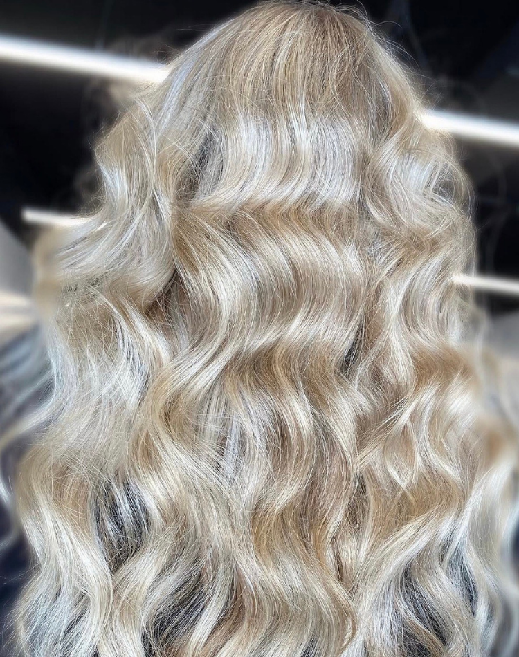 A hairdresser wearing black gloves applies light hair dye on a woman's blonde hair close to the scalp.