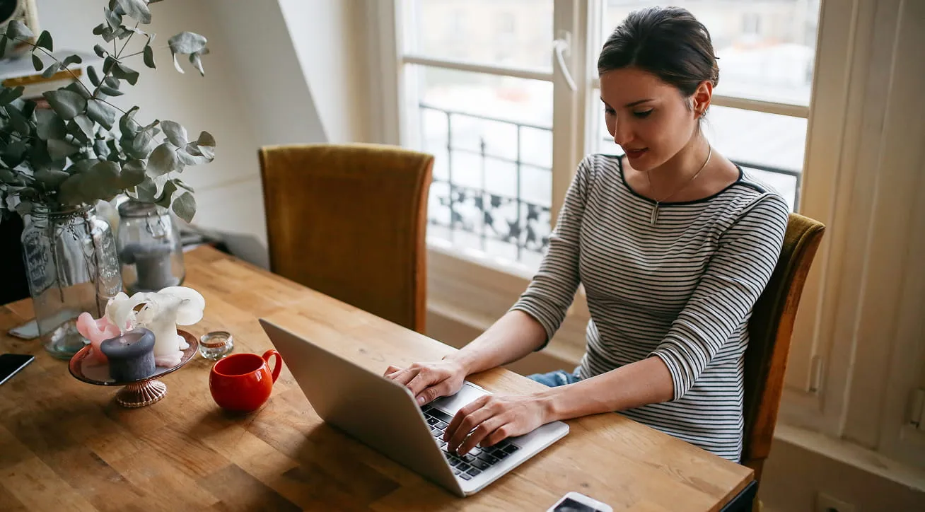 Person working on a laptop at a wooden table near a window with a coffee mug and plants.