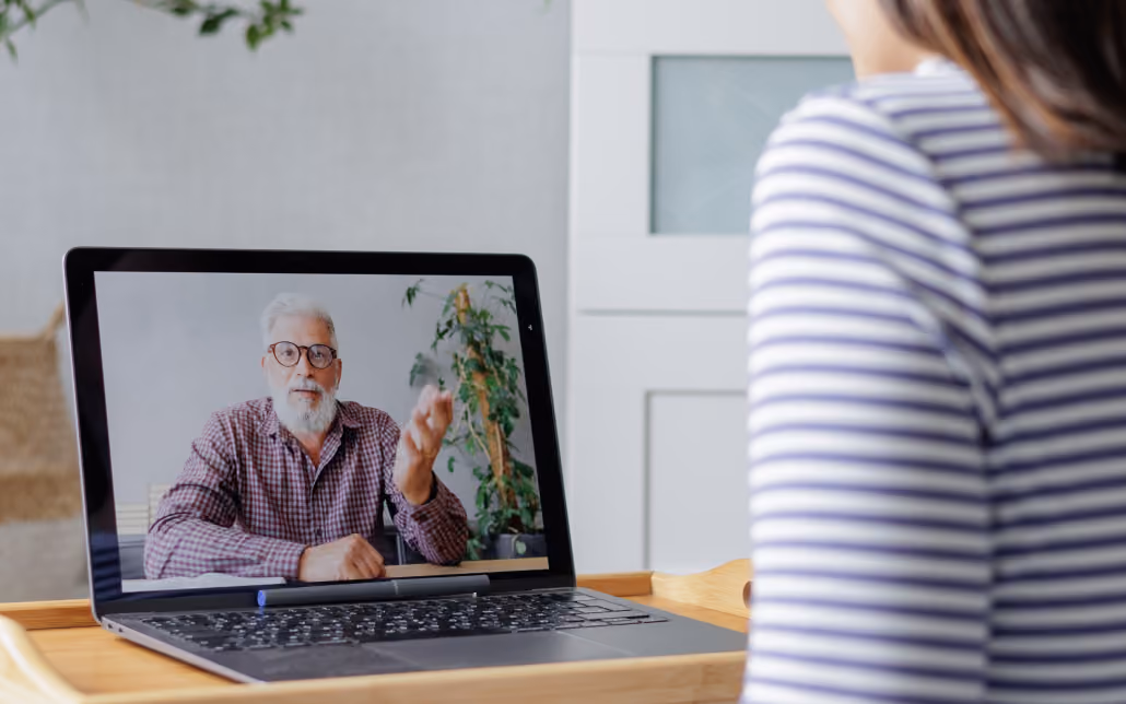 Person in striped shirt watching an older man with glasses speaking on a laptop screen.