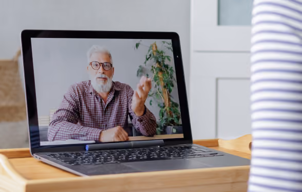 Older man with glasses speaking on a laptop screen during a video call at a wooden desk.