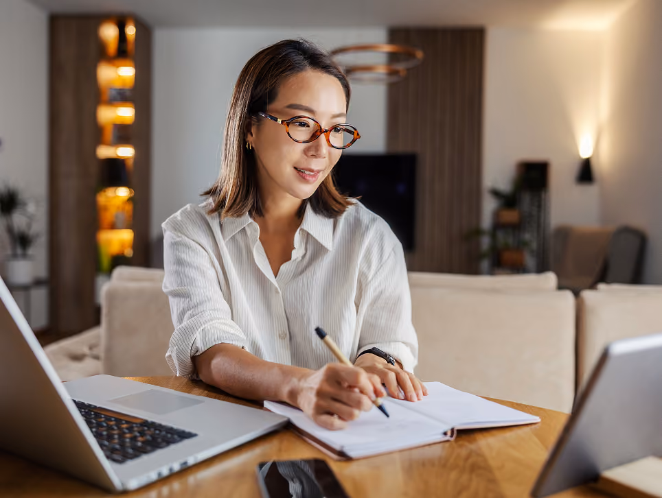 Woman with glasses writing in notebook at table with laptop and tablet in cozy living room.