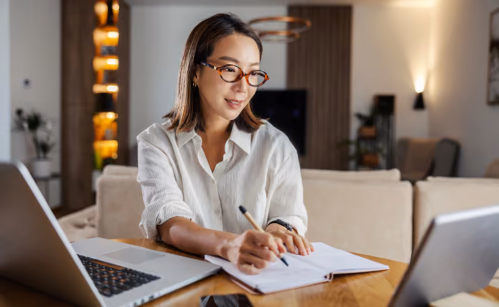 Woman with glasses writing in a notebook at a table with a laptop and tablet in a cozy room.