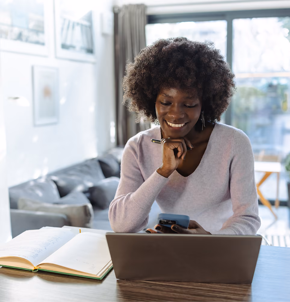 Smiling woman sitting at a table using a smartphone and laptop with an open book nearby in a bright room.