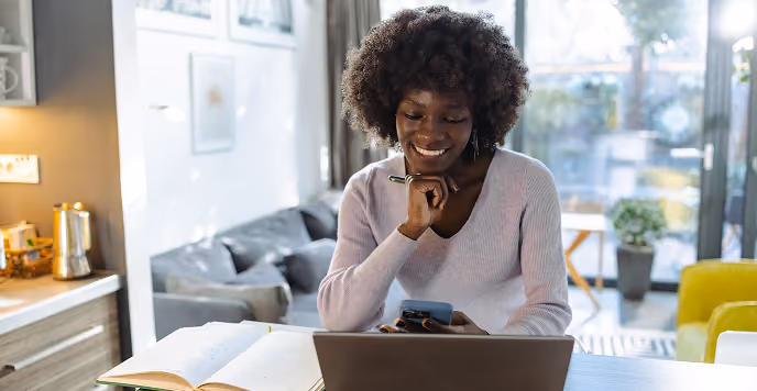 Smiling woman sitting at a table using a smartphone and laptop with an open book nearby in a bright room.