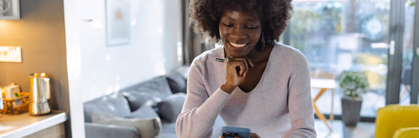 Smiling woman sitting at a table using a smartphone and laptop with an open book nearby in a bright room.