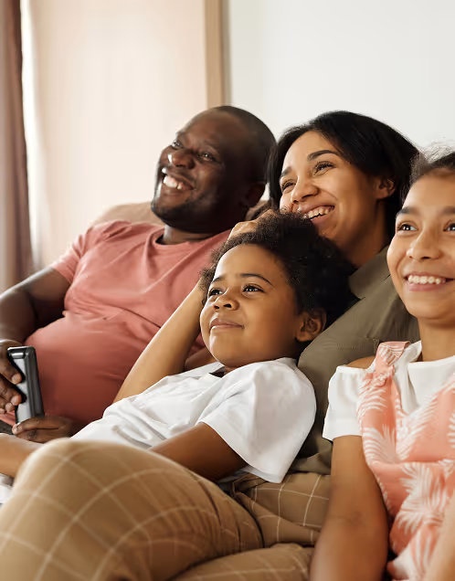 Smiling family of four sitting closely on a couch, watching TV together with a remote control in hand.