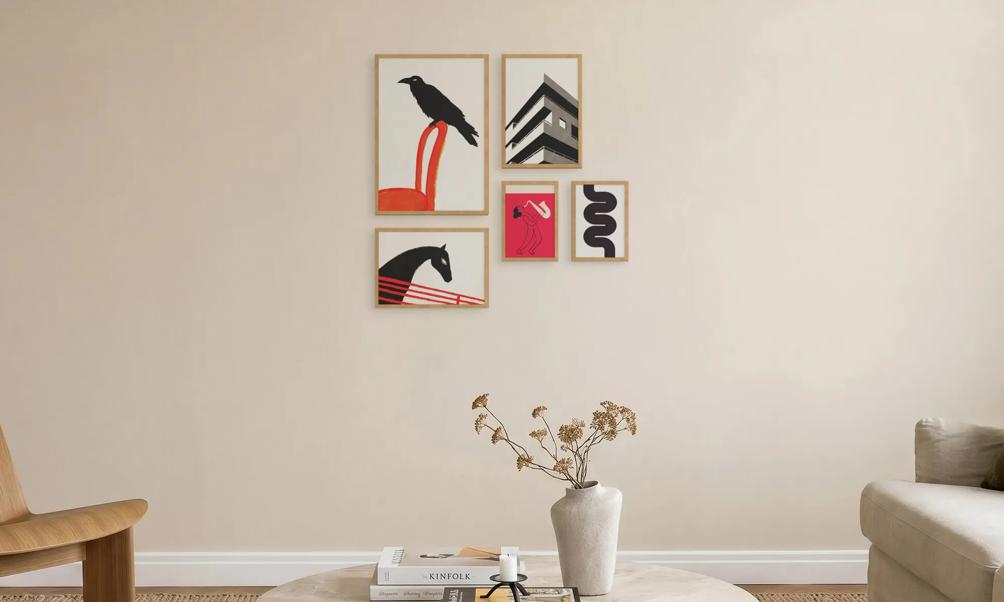 Minimalist living room wall with five framed art pieces: a black bird on a red chair, a black and white building, a red saxophone player, a black abstract curved shape, and a black horse with red lines on white background, above a table with dried flowers in a white vase, books, and a candle, flanked by a wooden chair and a beige sofa.