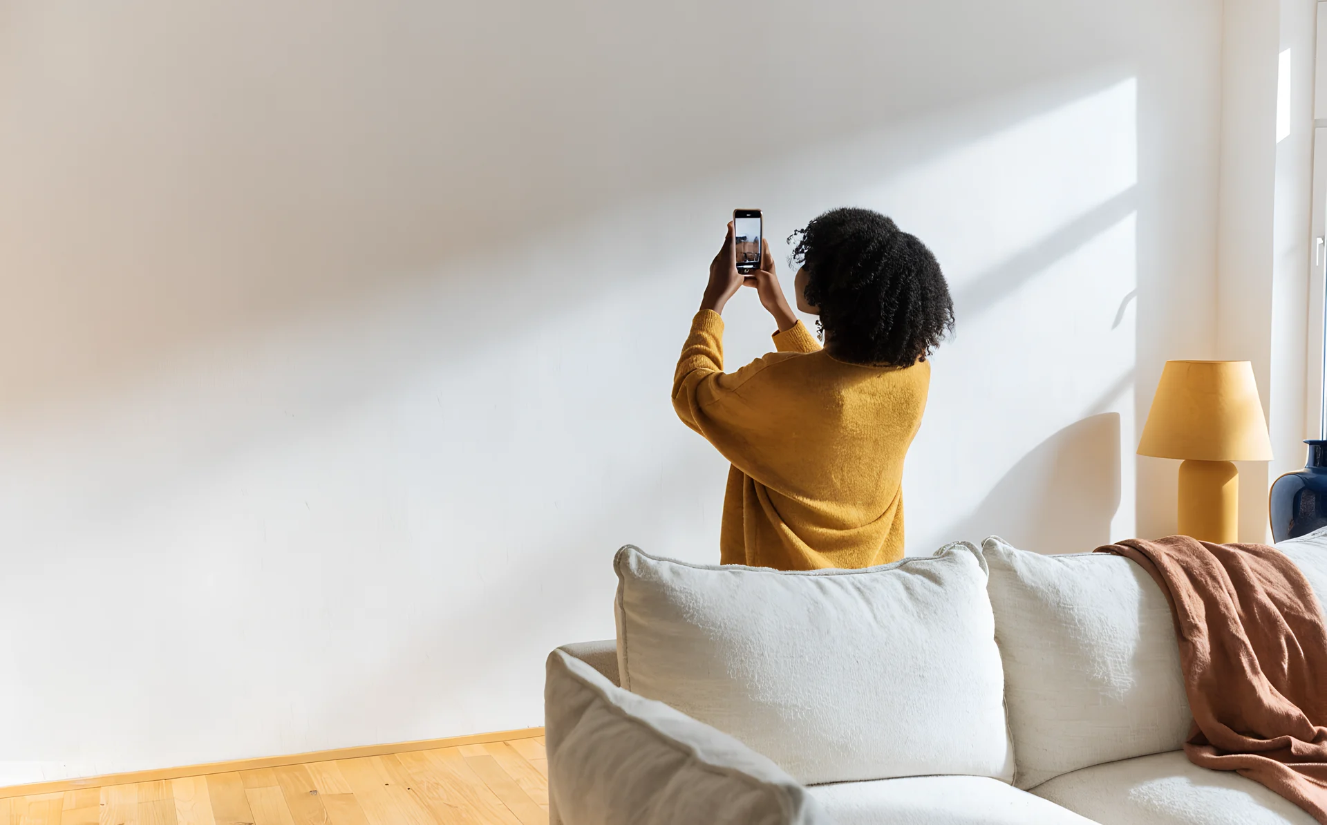 A woman with natural curls in a mustard jumper photographs an empty living room wall on her phone to plan a gallery wall