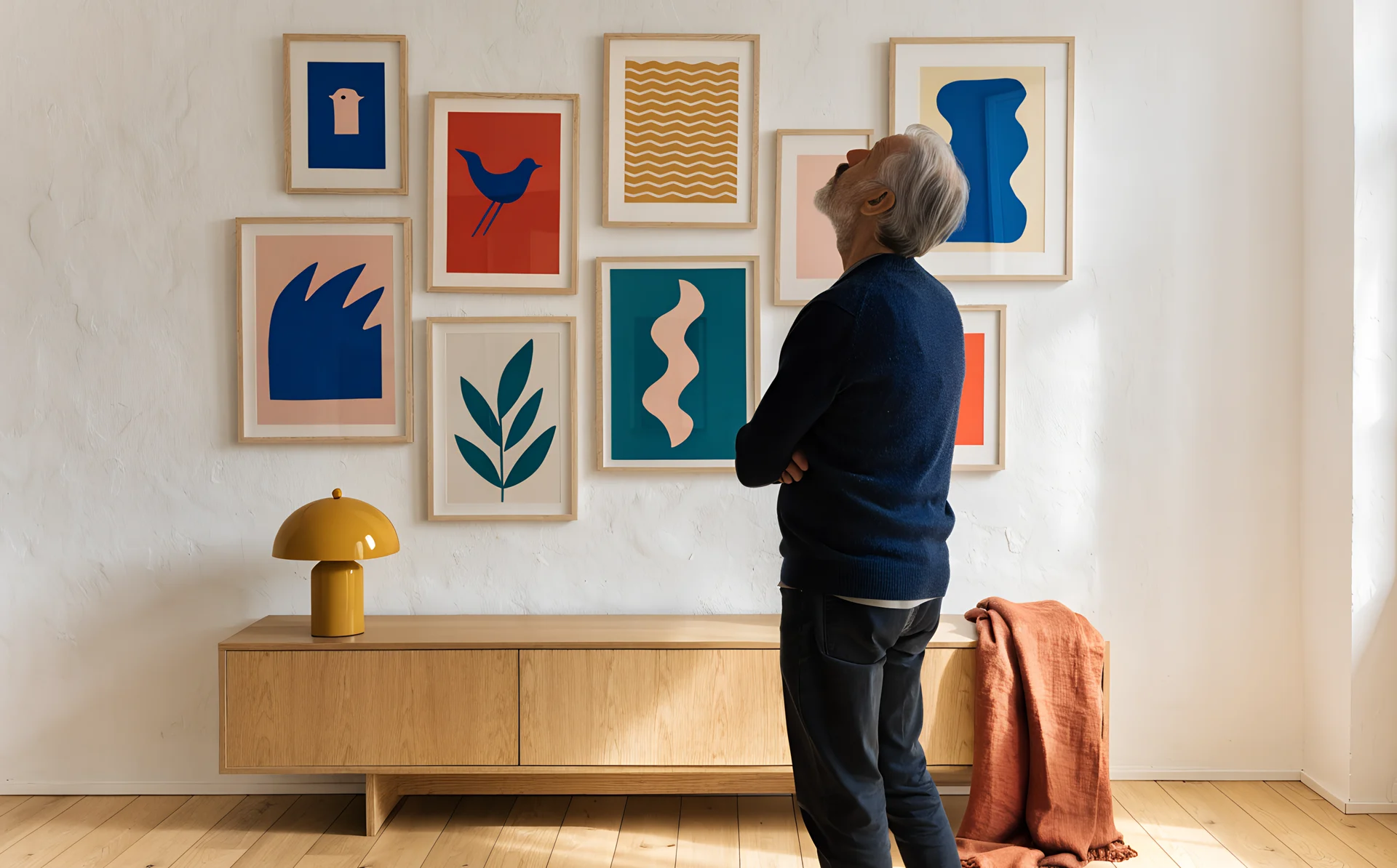 An older man in a navy cardigan stands back with his arms folded to judge the height of his gallery wall