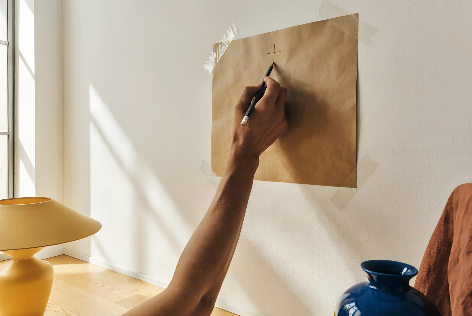 A hand marks the hanging point with a pencil on a paper template taped to the wall