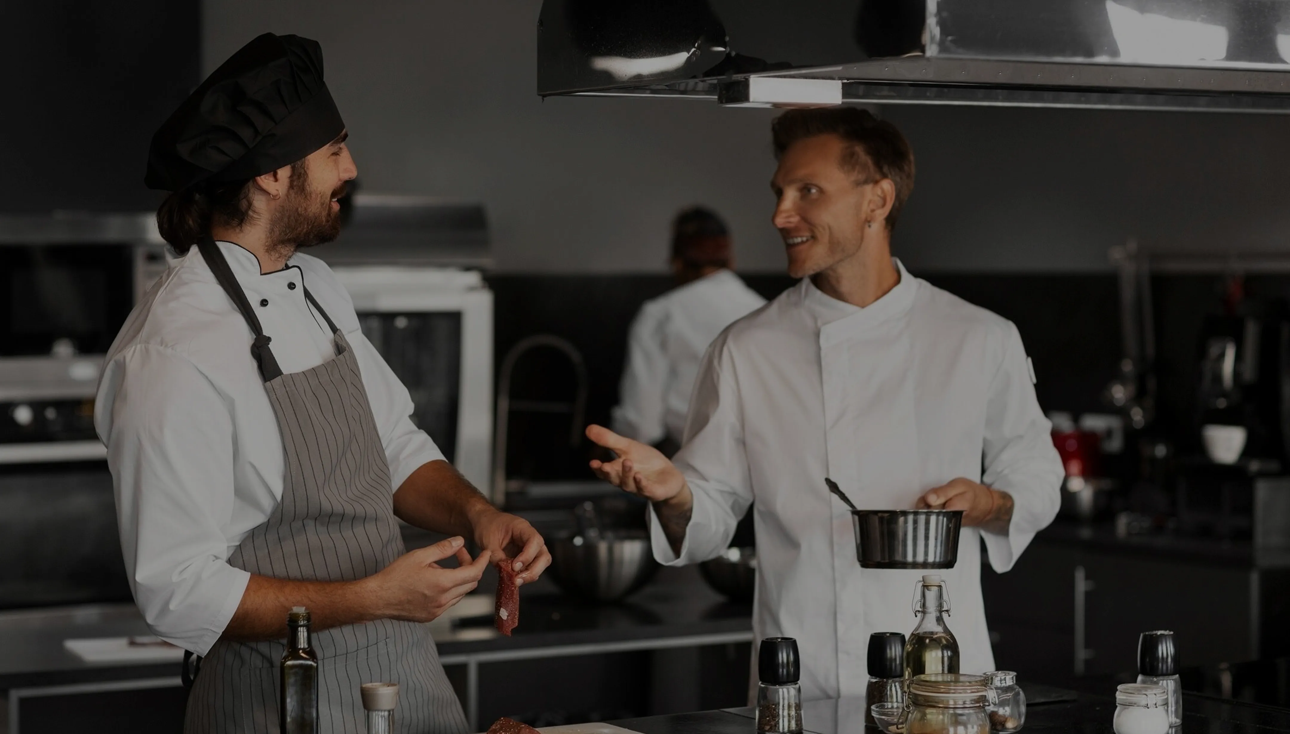 Two chefs talking in a kitchen, one holding a small pot and the other wearing an apron and hat.