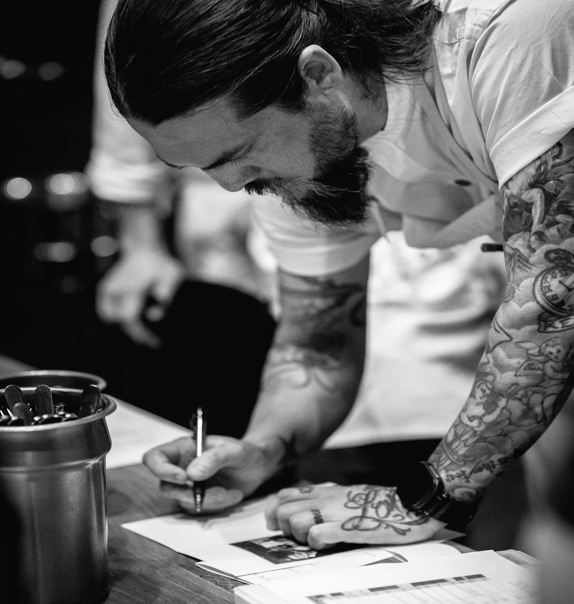 Tattooed man with beard and long hair writing on paper at a desk with a container of spoons nearby.