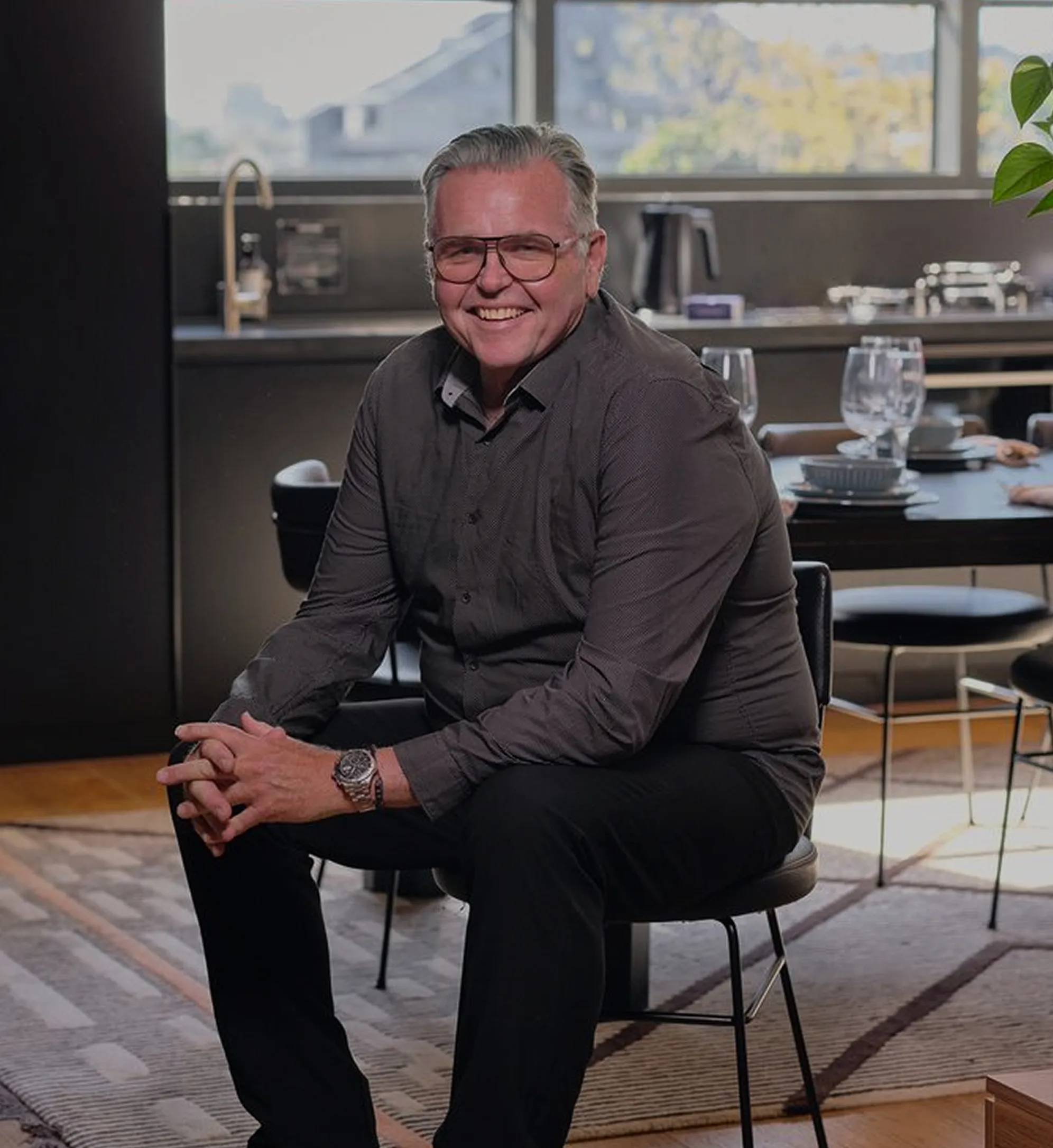 Smiling man wearing glasses and a dark shirt sitting on a chair in a modern dining room.