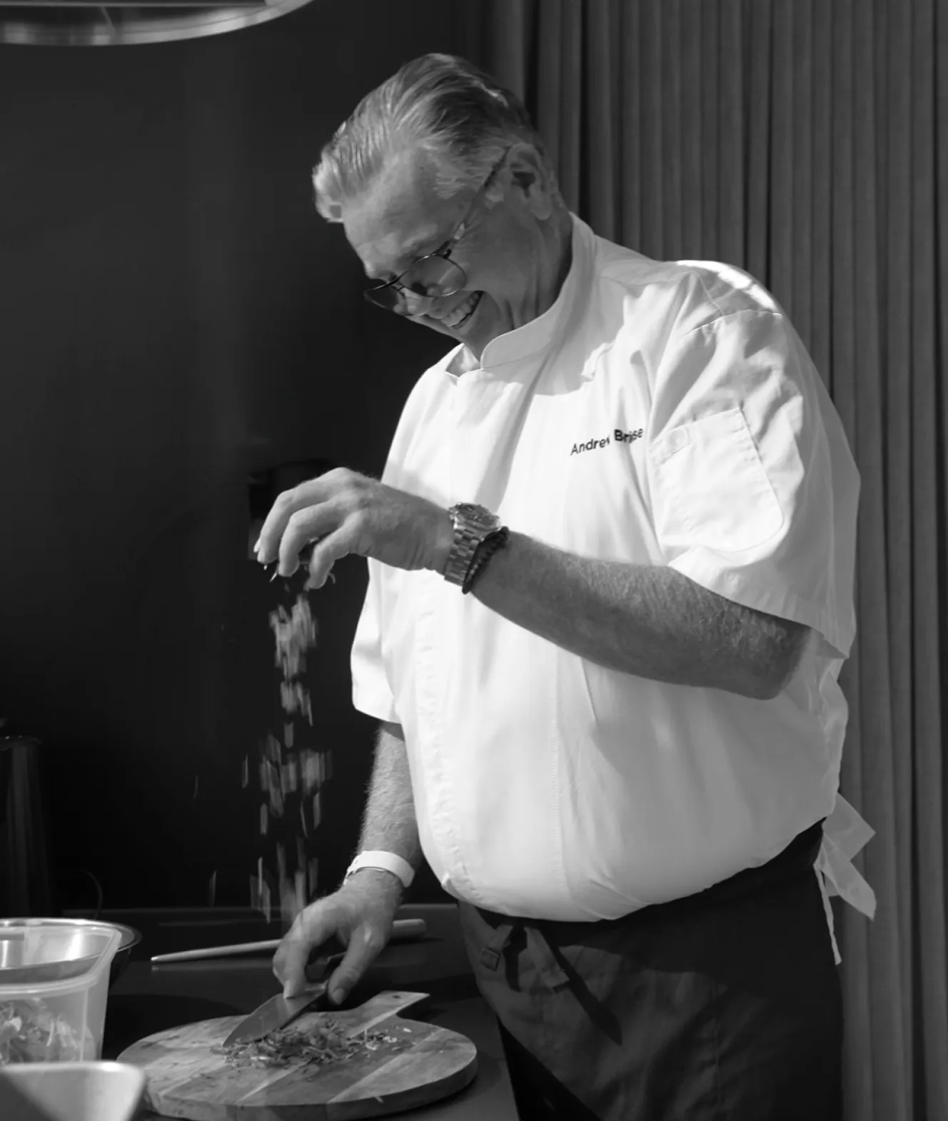 Male chef smiling while sprinkling seasoning over chopped ingredients on a wooden cutting board.