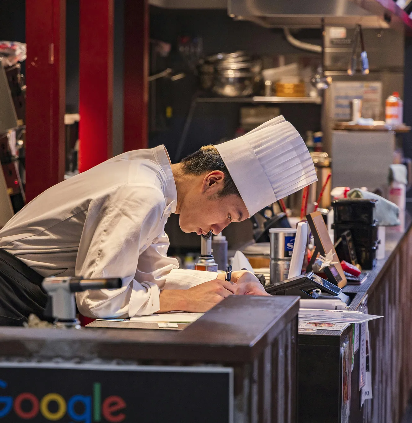 Chef in white uniform and hat writing on paper at a restaurant counter with kitchen tools nearby.