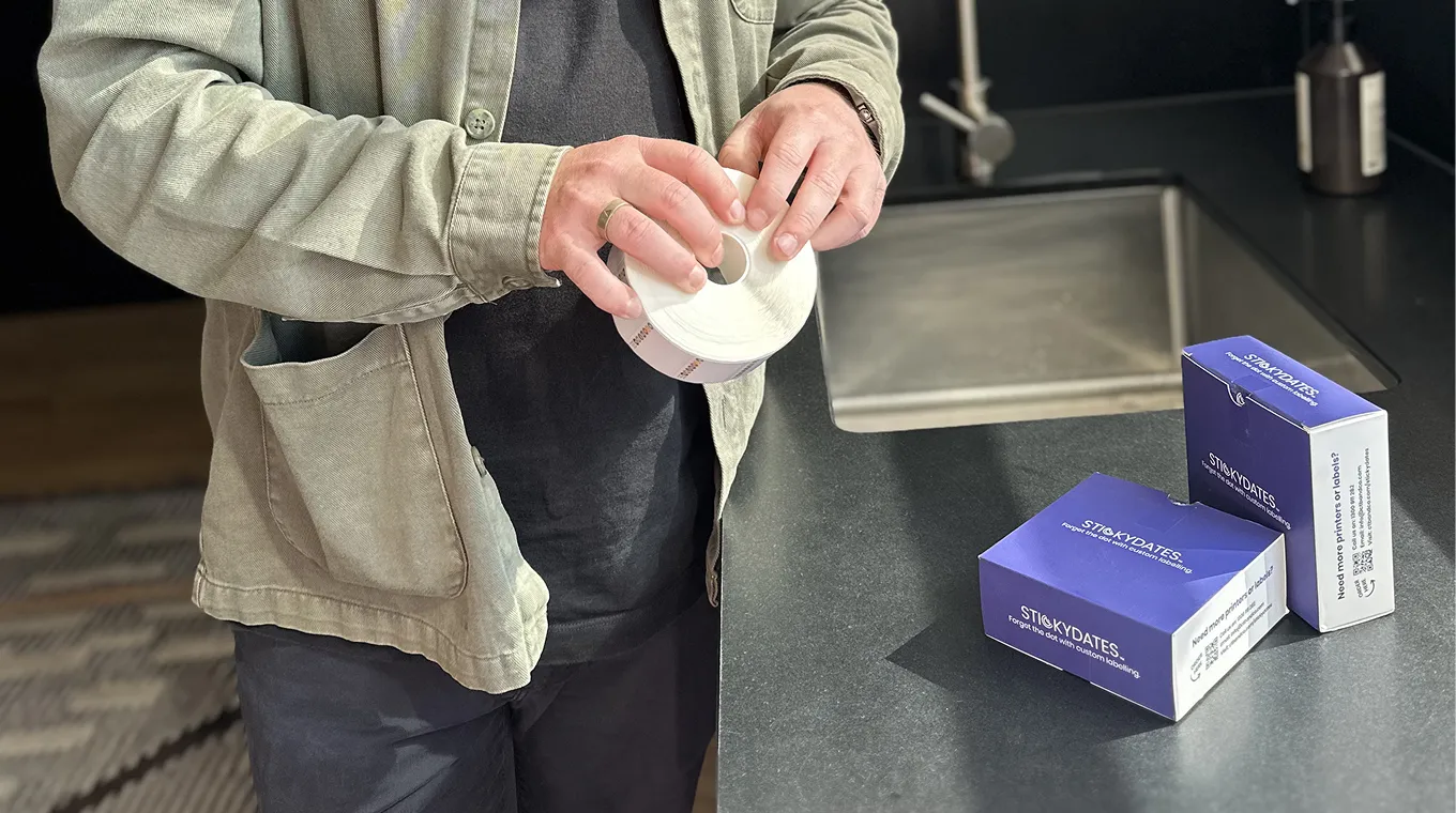 Person holding a roll of blank labels beside two purple Stickydates boxes on a dark countertop.