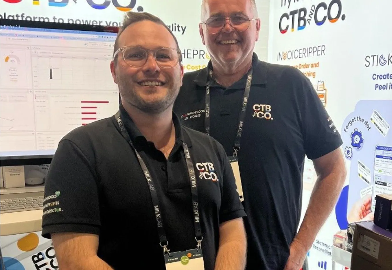 Two men smiling wearing black CTB and CO. polo shirts at a trade booth with computer screens behind them.