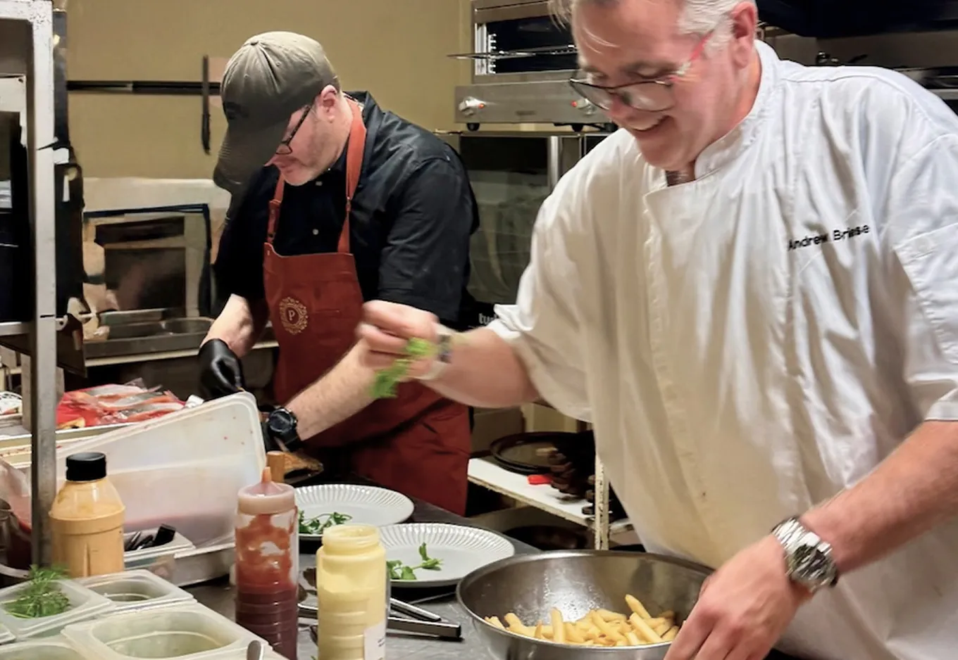 Two chefs preparing plates in a kitchen, one adding greens, the other focused on food prep.