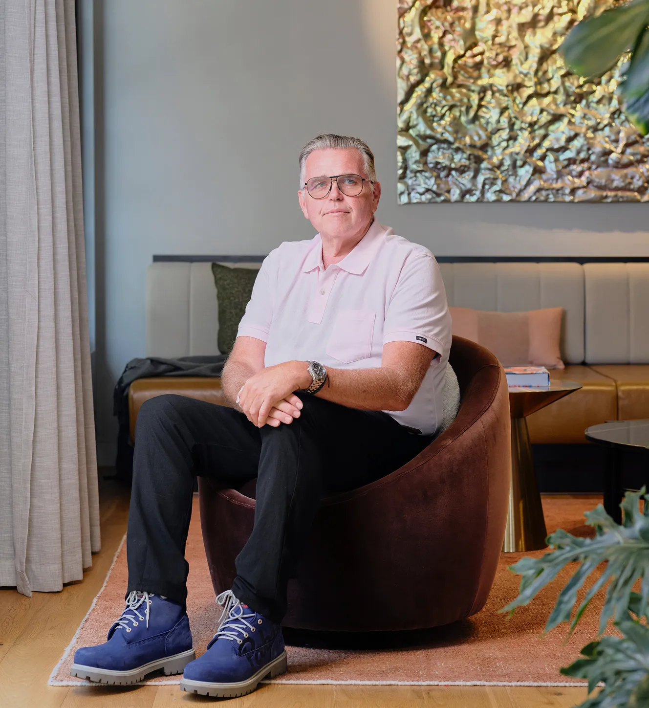 Man with gray hair and glasses sitting in a brown chair wearing a light pink polo and blue boots.