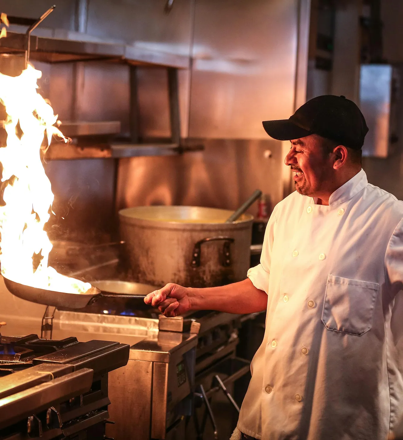 Chef in white coat and black cap flipping food on a frying pan with flames in a commercial kitchen.