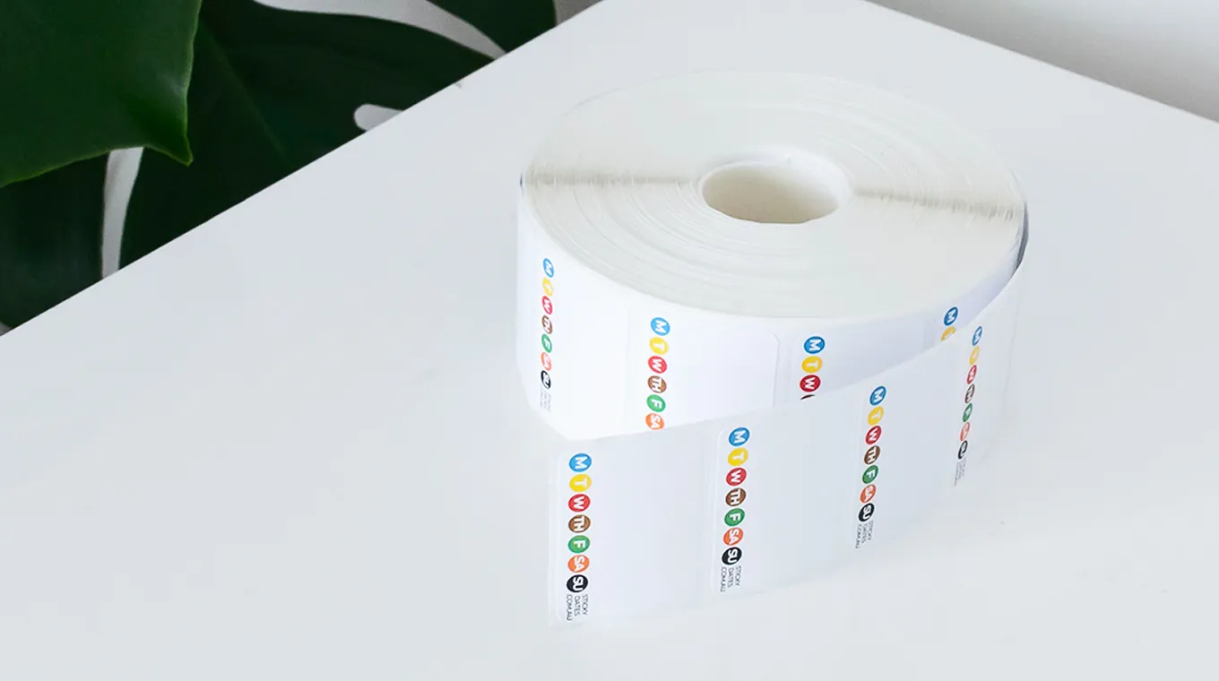 A roll of white labels with colorful icons and text placed on a white surface near green leaves.