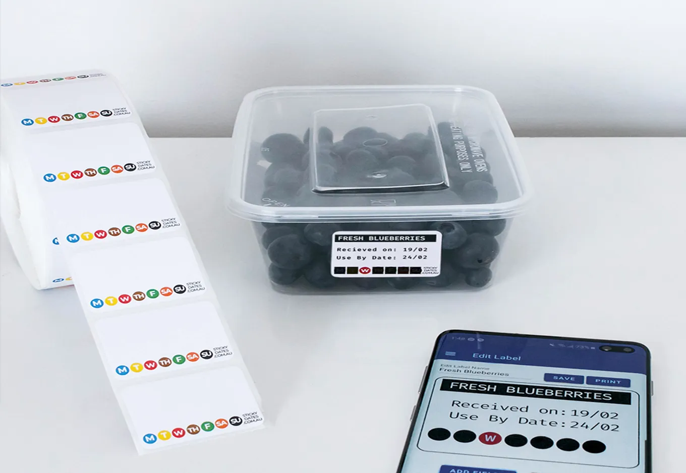 Plastic container of blueberries with tracking label beside label stickers and smartphone showing label editing.