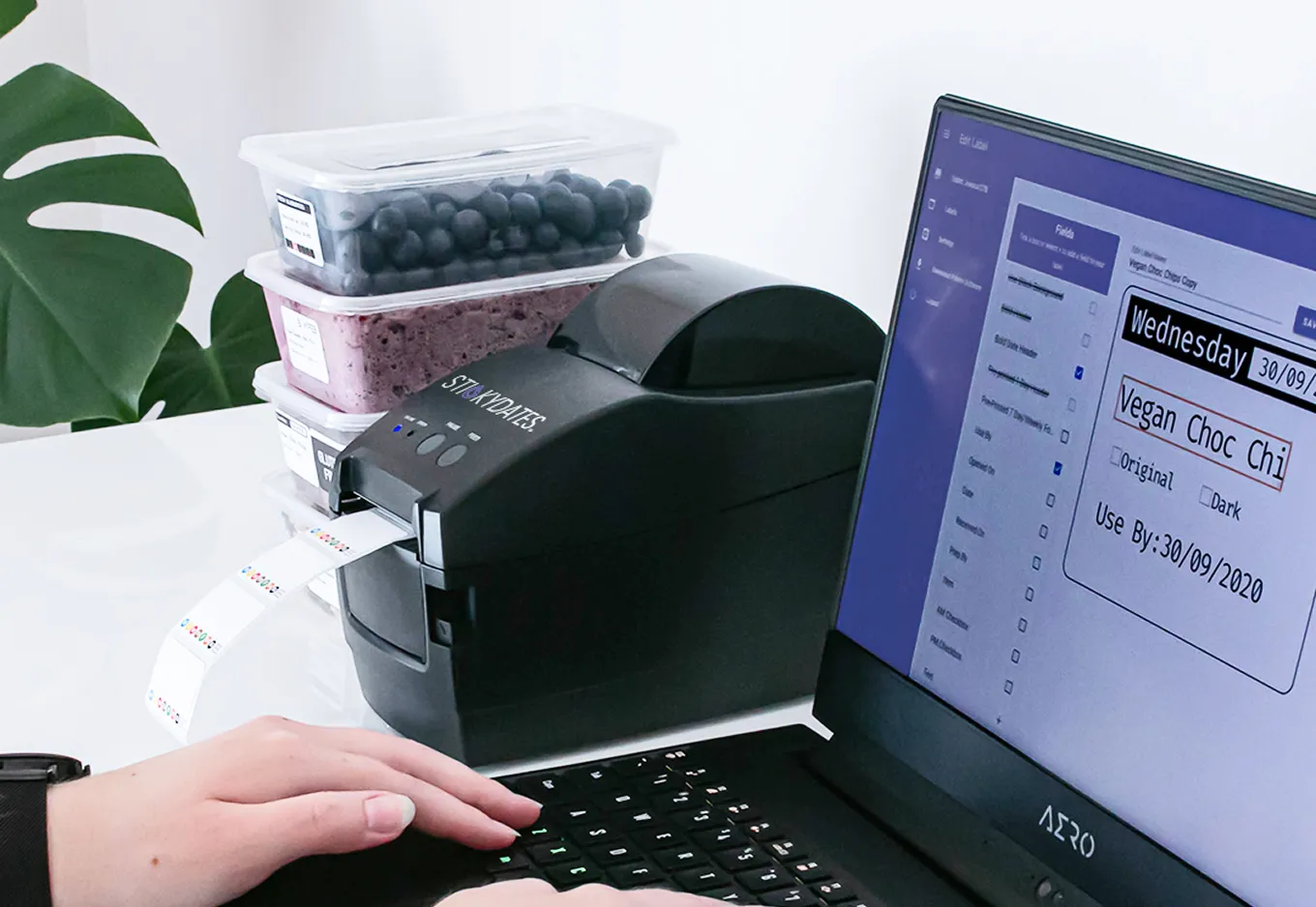 Person typing on laptop next to label printer printing food labels, with stacked food containers and plant nearby.