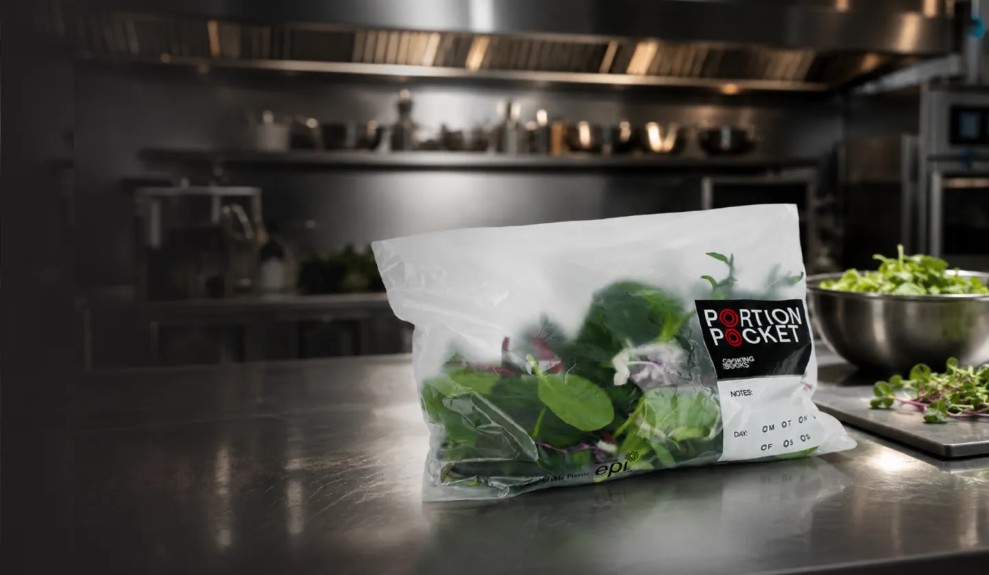Plastic bag labeled Portion Pocket containing mixed leafy greens on a stainless steel kitchen counter.