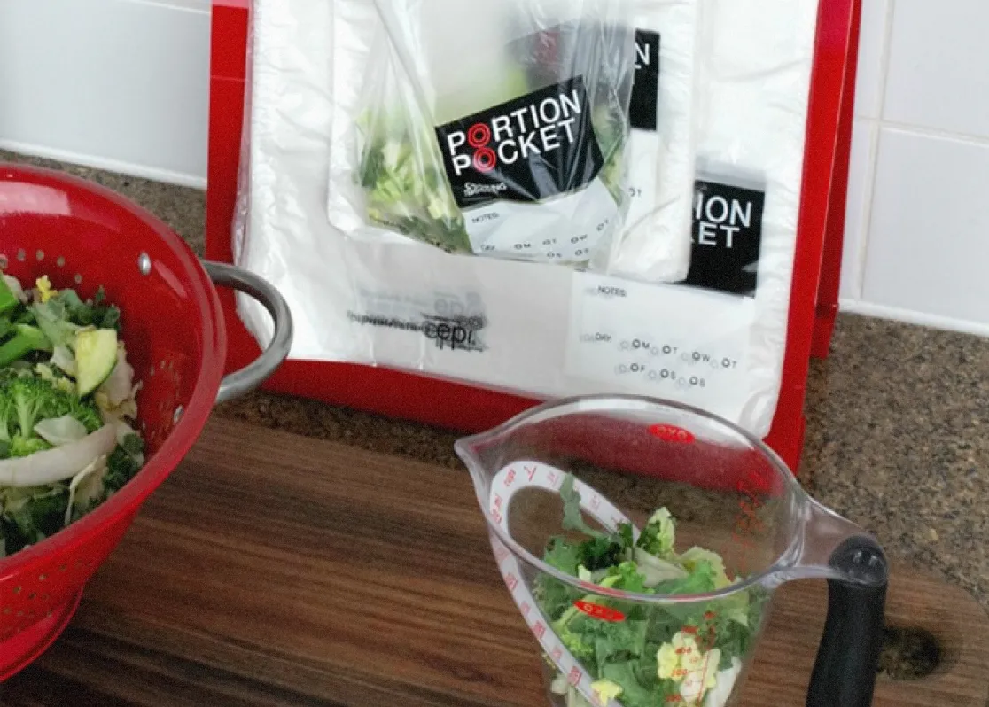 Red colander with green vegetables beside a measuring cup and Portion Pocket bags on a kitchen counter.