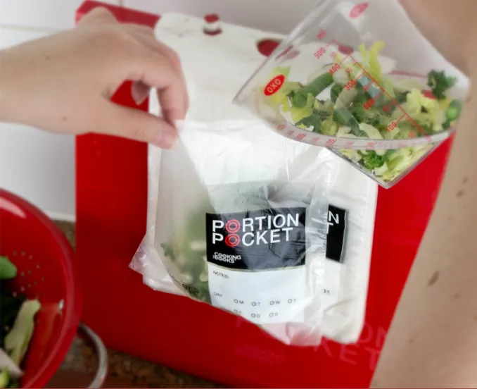 Hand filling a Portion Pocket bag with chopped vegetables from a measuring cup in a kitchen.