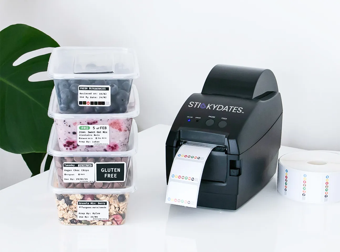 Stacked food containers with labels next to a black STICKYDATES label printer on a white surface.