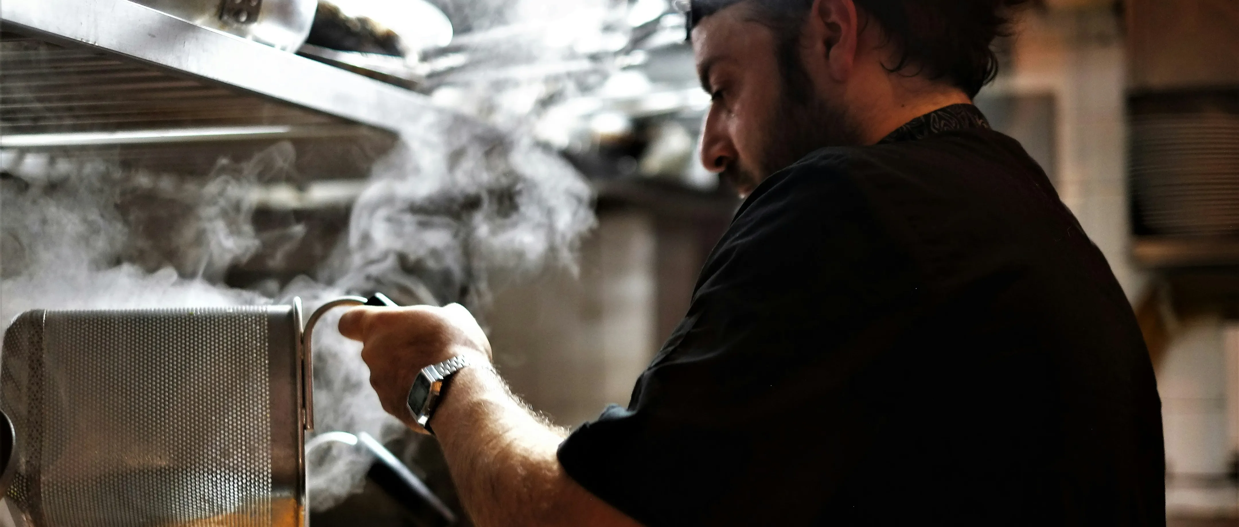 Man in black shirt cooking with steam rising inside a kitchen.