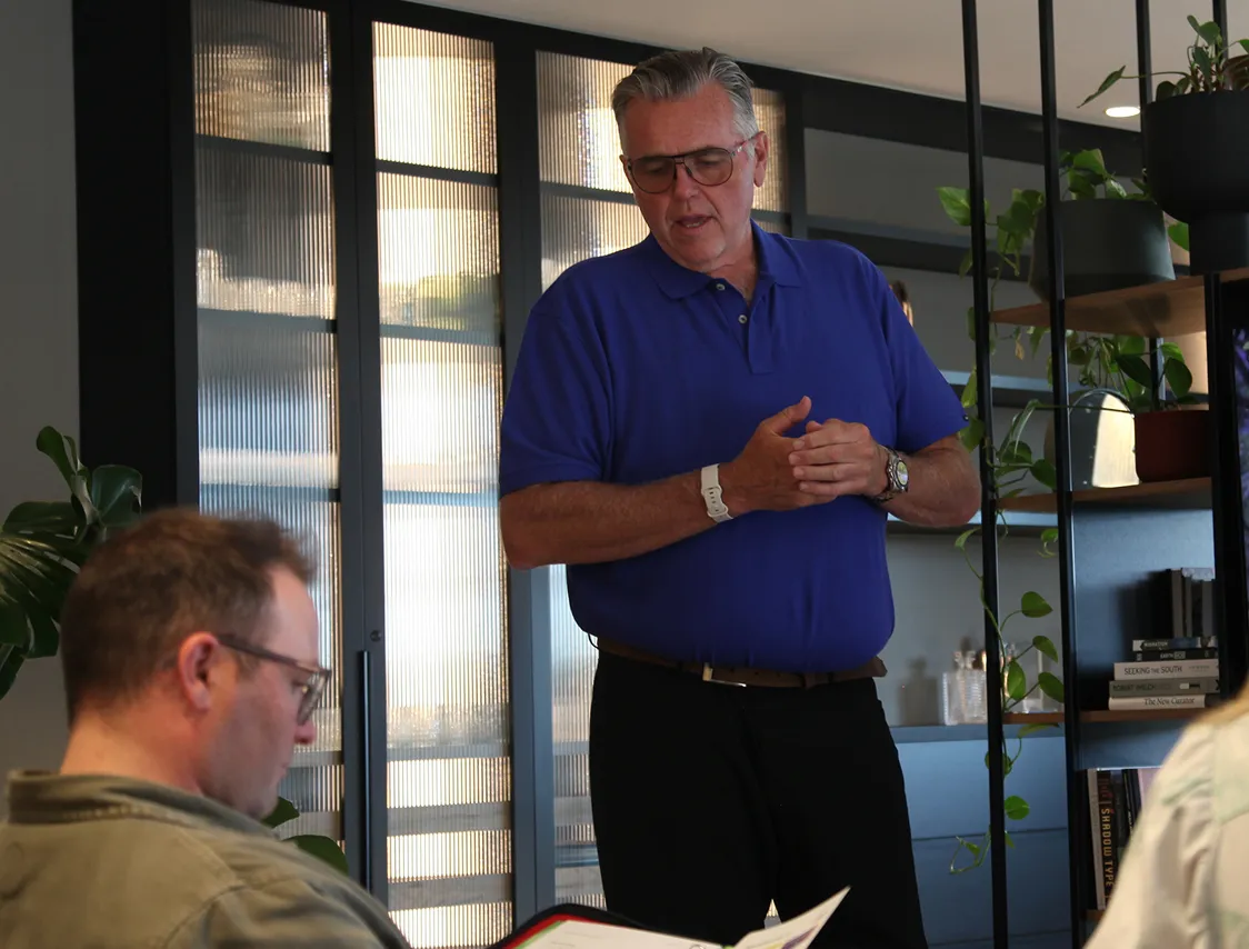 Man in a blue polo shirt speaking to seated people in a modern office space with plants and shelves.