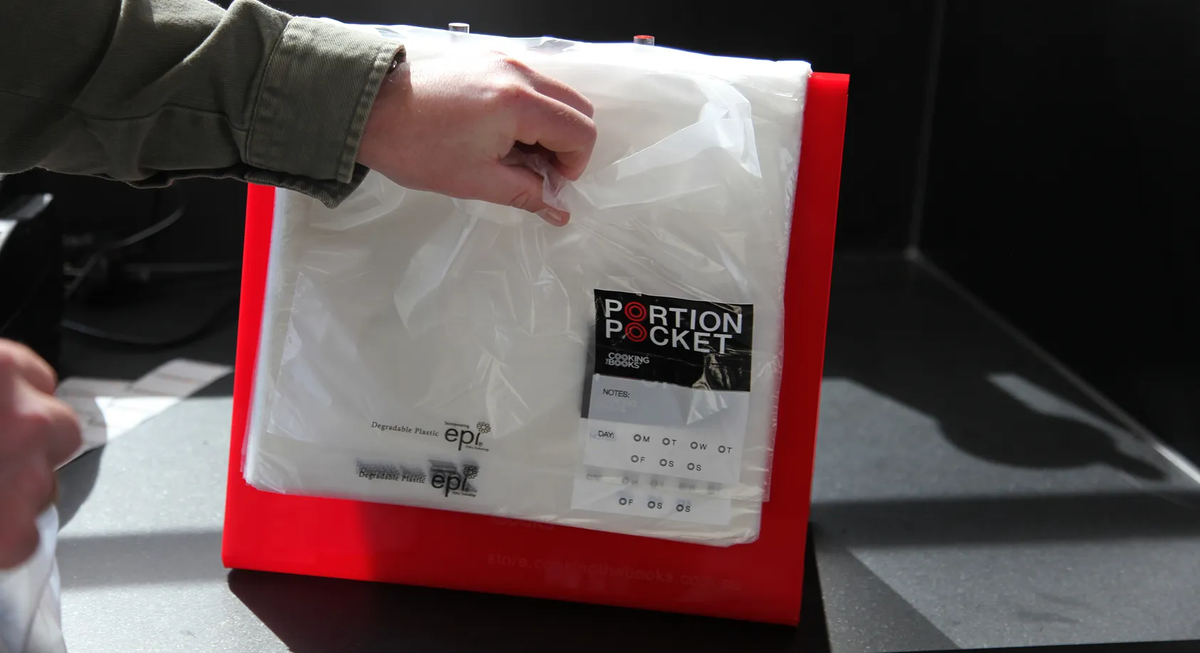 Hand grabbing a clear plastic bag labeled Portion Pocket against a red stand on a gray surface.