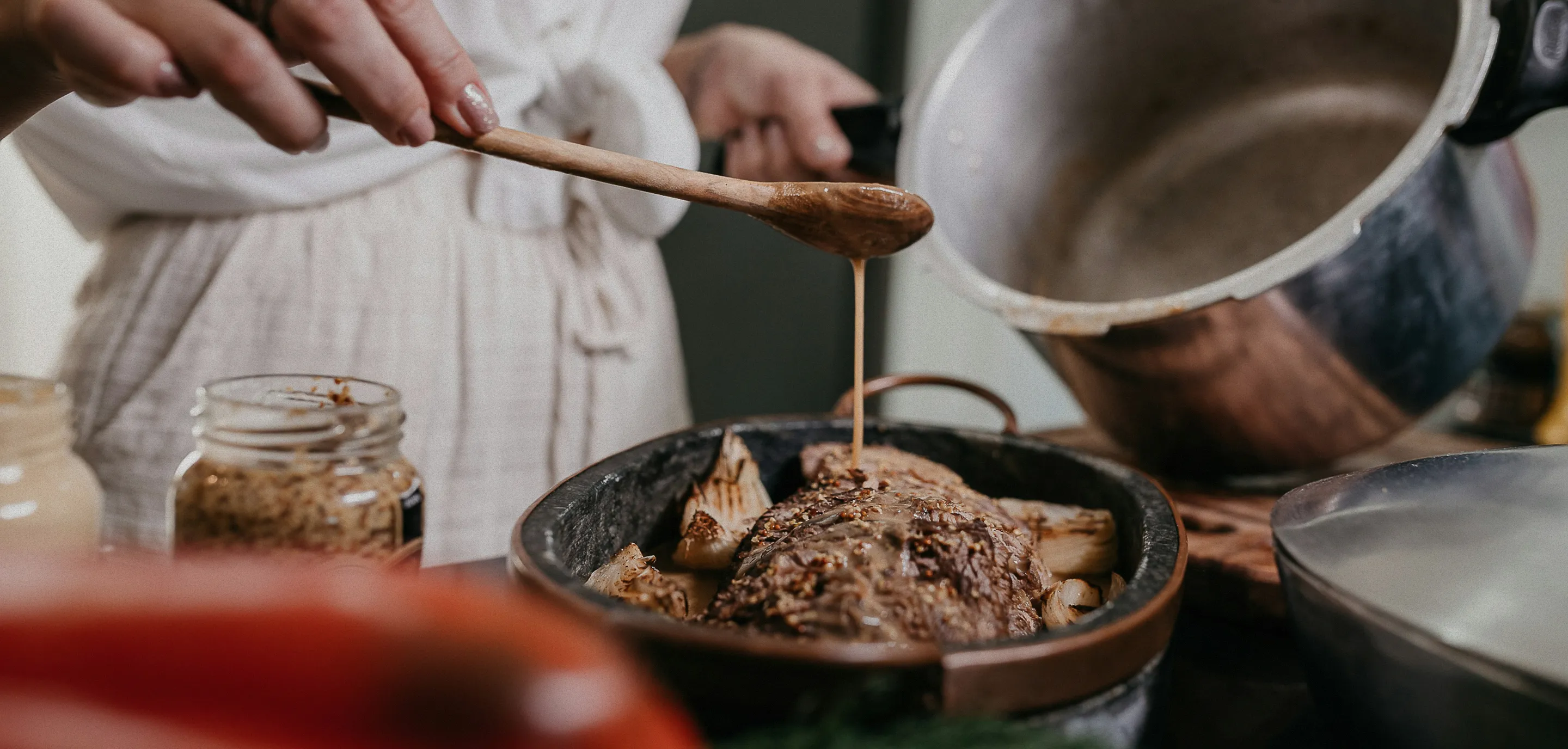 Person drizzling sauce from a wooden spoon onto cooked meat in a skillet with vegetables.