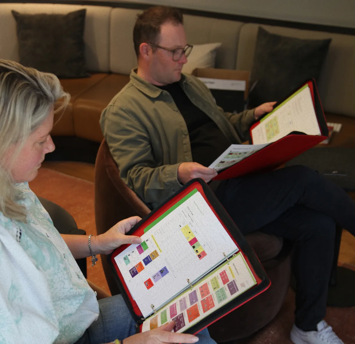 Two people sitting on chairs in a living room, reviewing colorful charts in red binders.