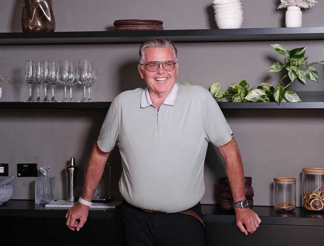 Smiling older man with glasses leaning on a black countertop in a kitchen with shelves behind him.