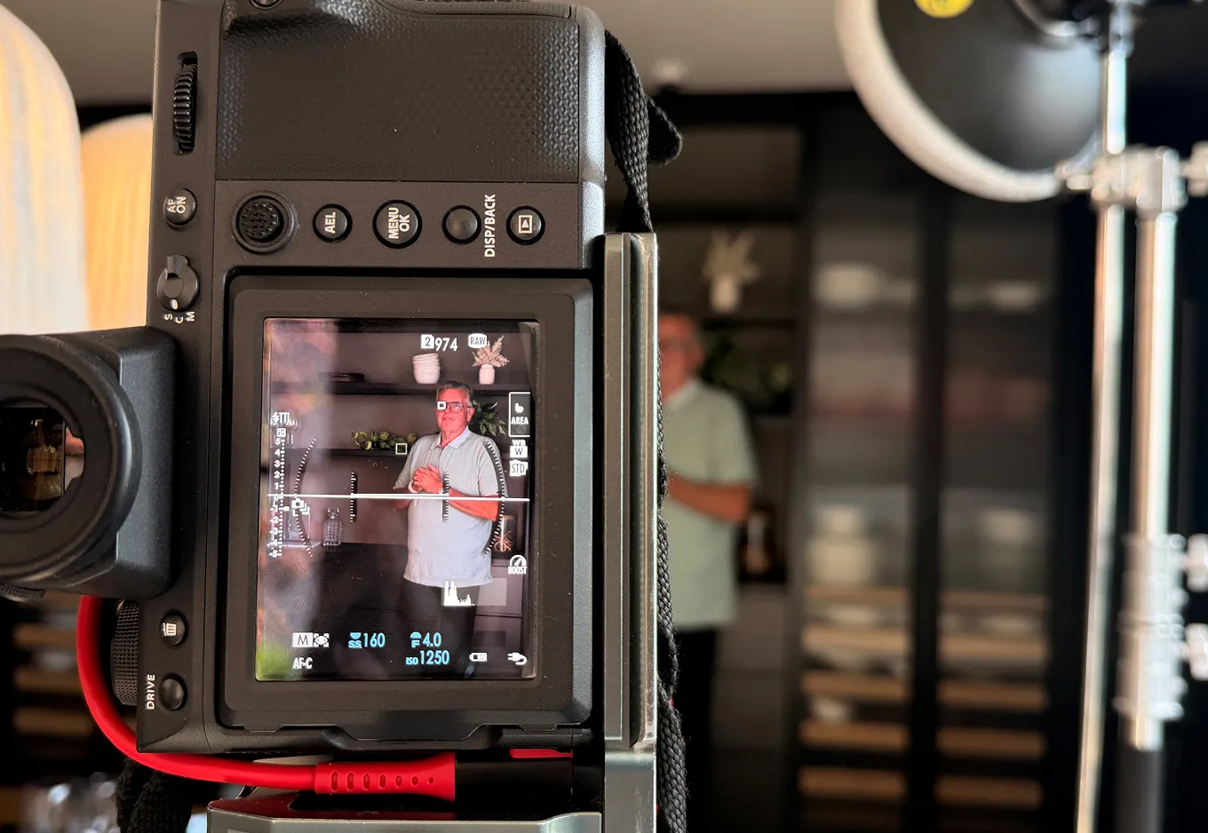 Camera screen showing a man in glasses posing indoors with shelves and decor in the background.