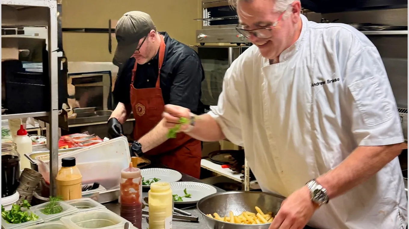 Two chefs prepare food in a kitchen with plates, sauces, and a bowl of fries.