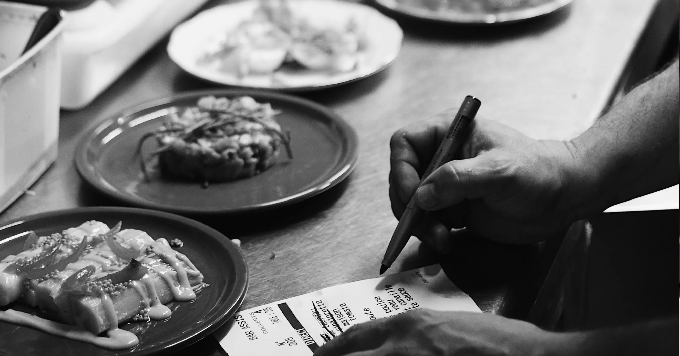 Hand holding pen over order ticket with plated dishes on a kitchen counter in black and white.